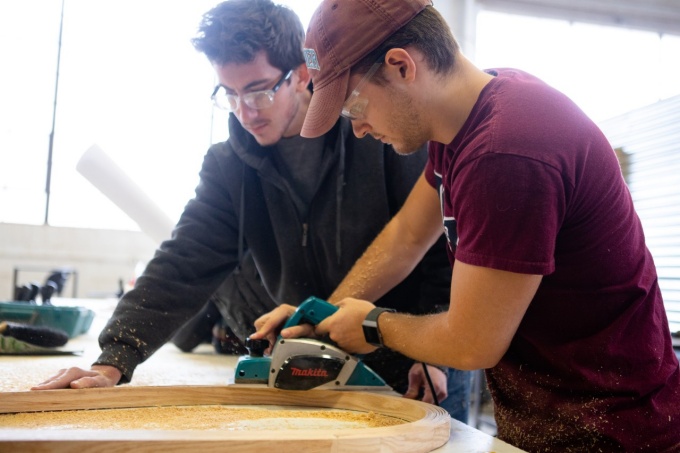 Students wear protective glasses and receive instruction while operating power tools. 