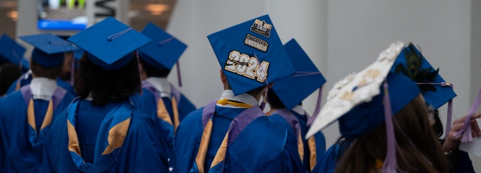 The photograph captures a group of graduates wearing blue caps and gowns. These graduation caps are adorned with various designs, texts, and symbols. The graduates face away from the camera, waiting for a commencement ceremony to begin. 