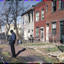A Black man stands along the street in front of a row of dilapidated buildings. 