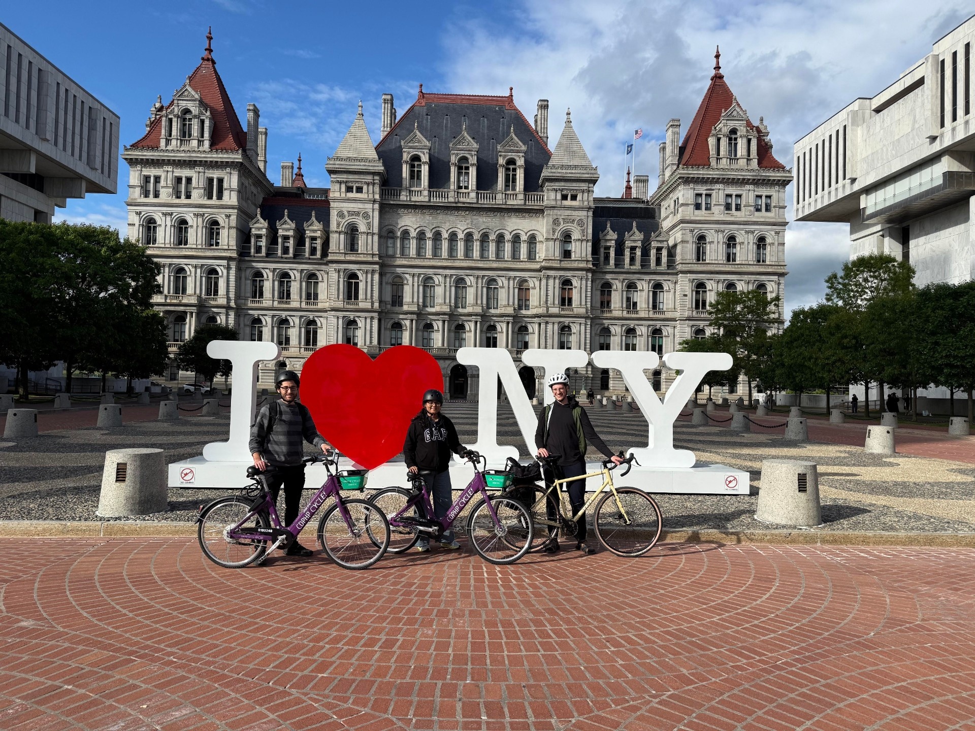 Three SMI employees stand next to bikes in front of the "I love New York" sign with the state capital building in the background.